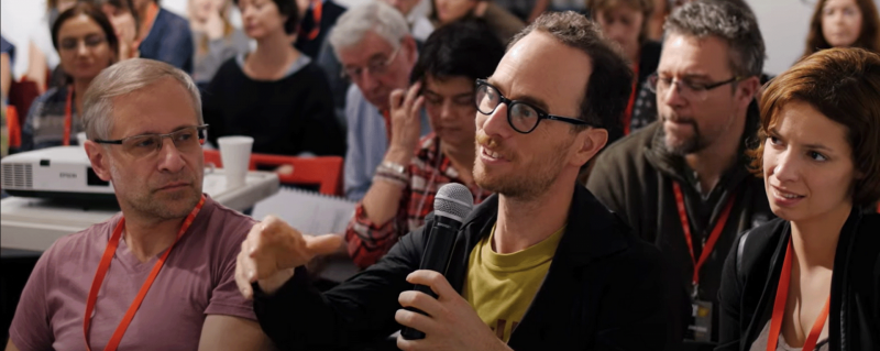 W sali konferencyjnej, podczas spotkania mężczyzna mówi do mikrofonu. 